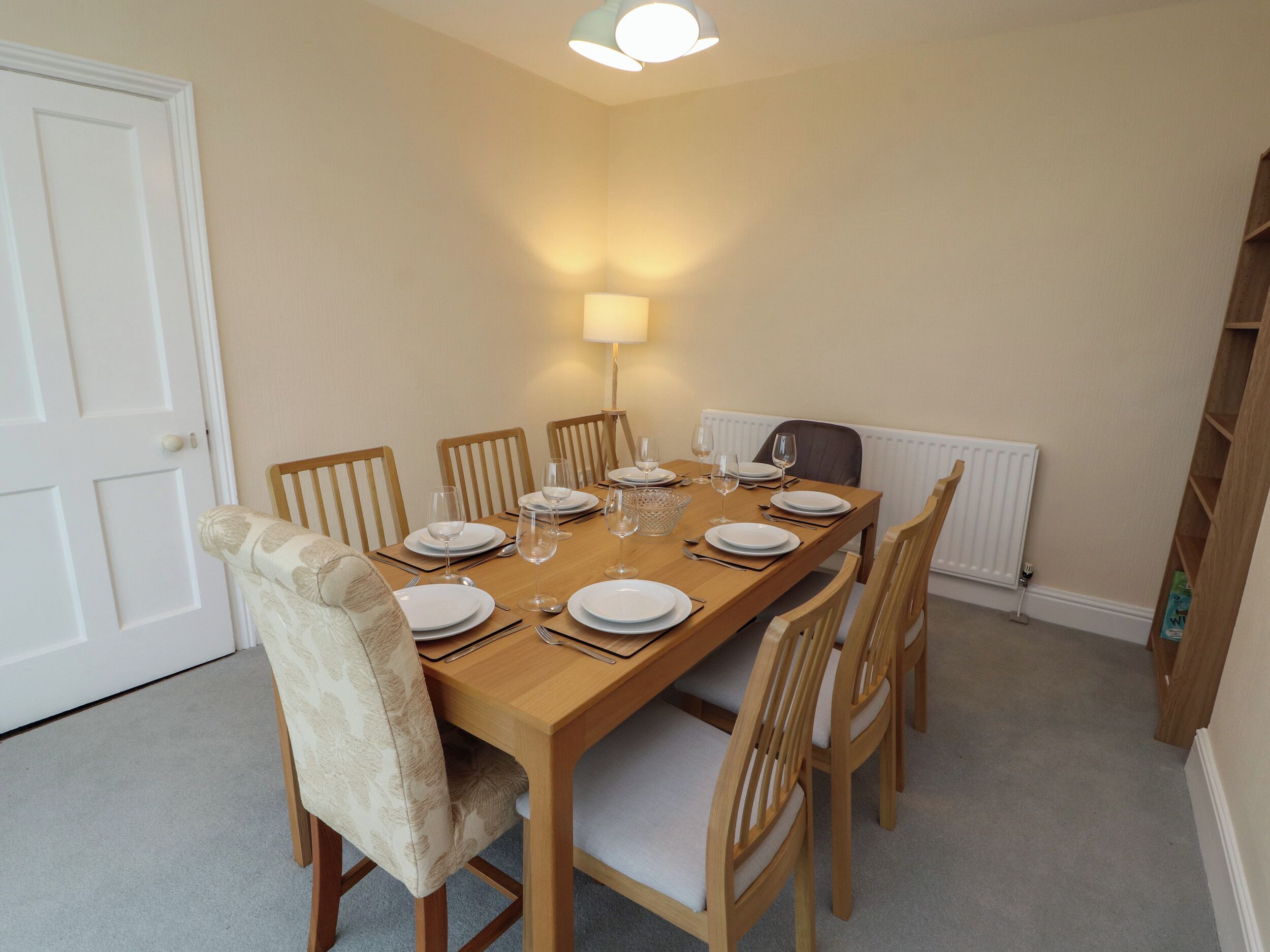 A dining room with a wooden table set for six, surrounded by six wooden chairs with cushioned seats. A white radiator is visible on the wall, and a white door is on the left.
