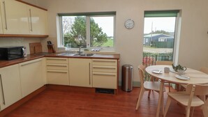 A kitchen with wooden cabinets, a sink, and a window. There is a dining table with chairs and a clock on the wall. - Burnbank (Maryport)
