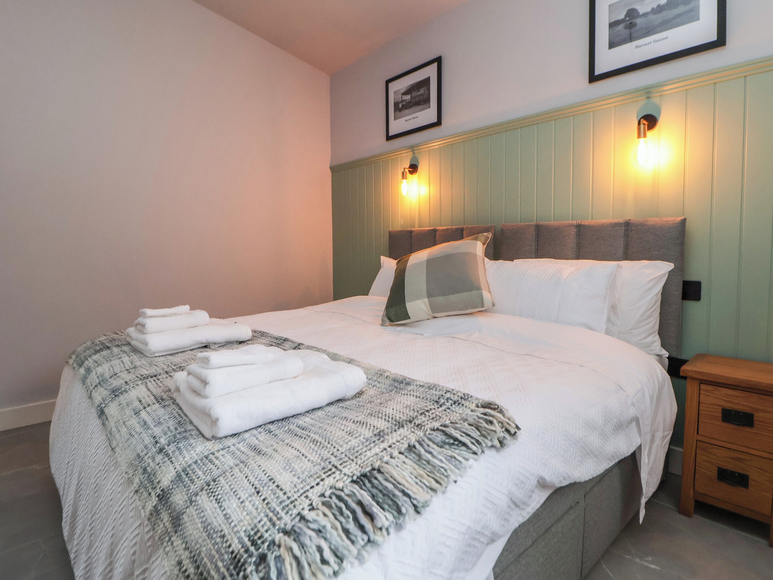 Een net opgemaakt bed met witte lakens en een geruit plaid, een nachtkastje met een lamp, en een lijst met een foto aan de muur.