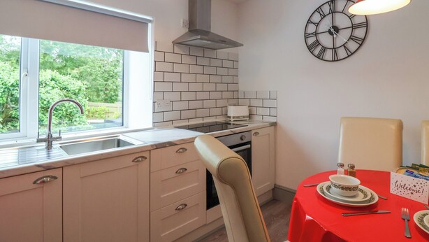 A modern kitchen with a window, a red table set for dining, and a clock on the wall. - Cosy Nest (Glastonbury)