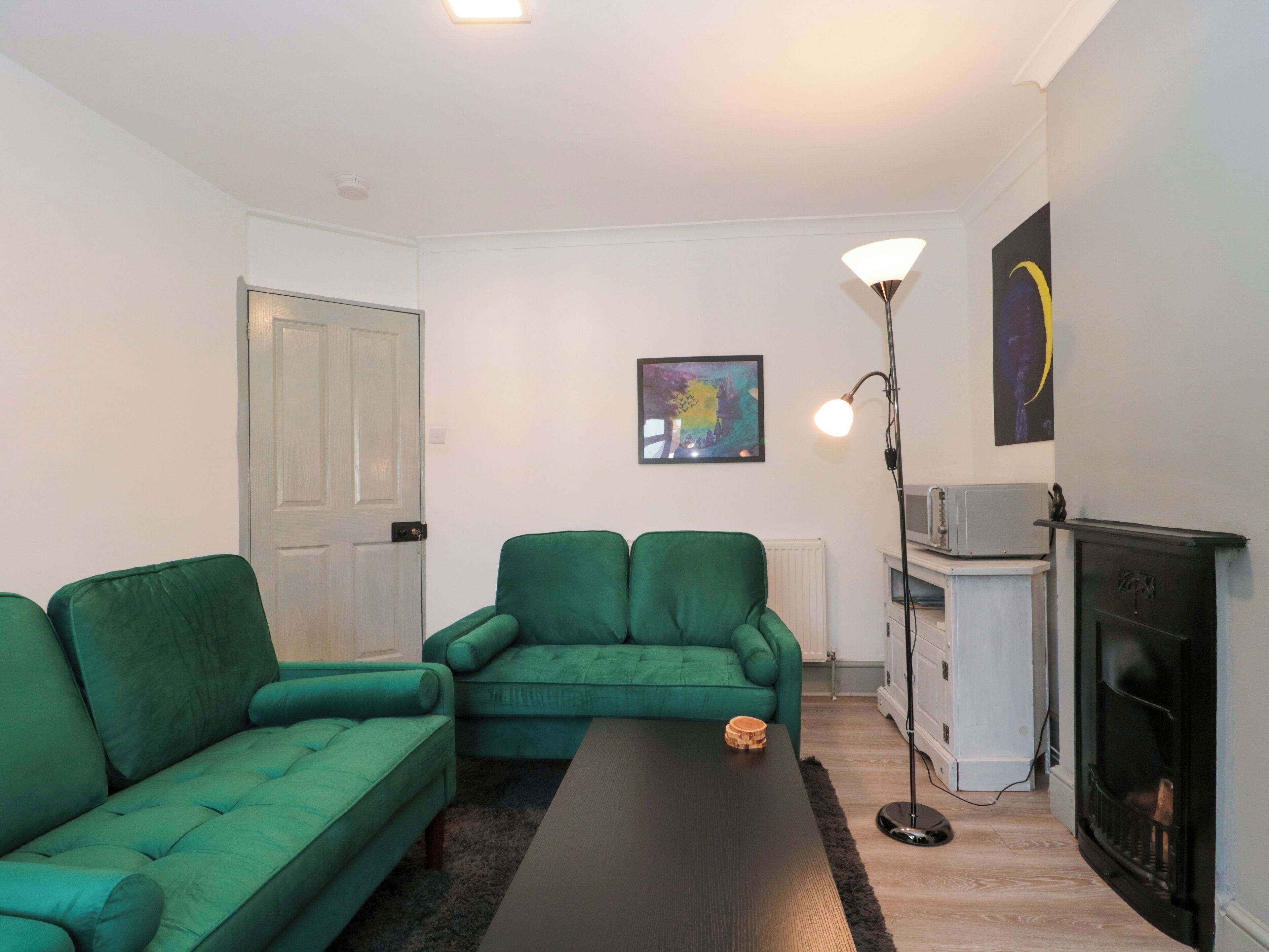 A living room with a green sofa set, a black coffee table, a fireplace, a floor lamp, and a framed abstract painting.