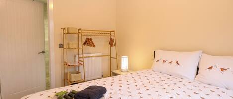 A neatly made bed with a bird pattern, a wooden drying rack with baskets, and a bedside table with a lamp.