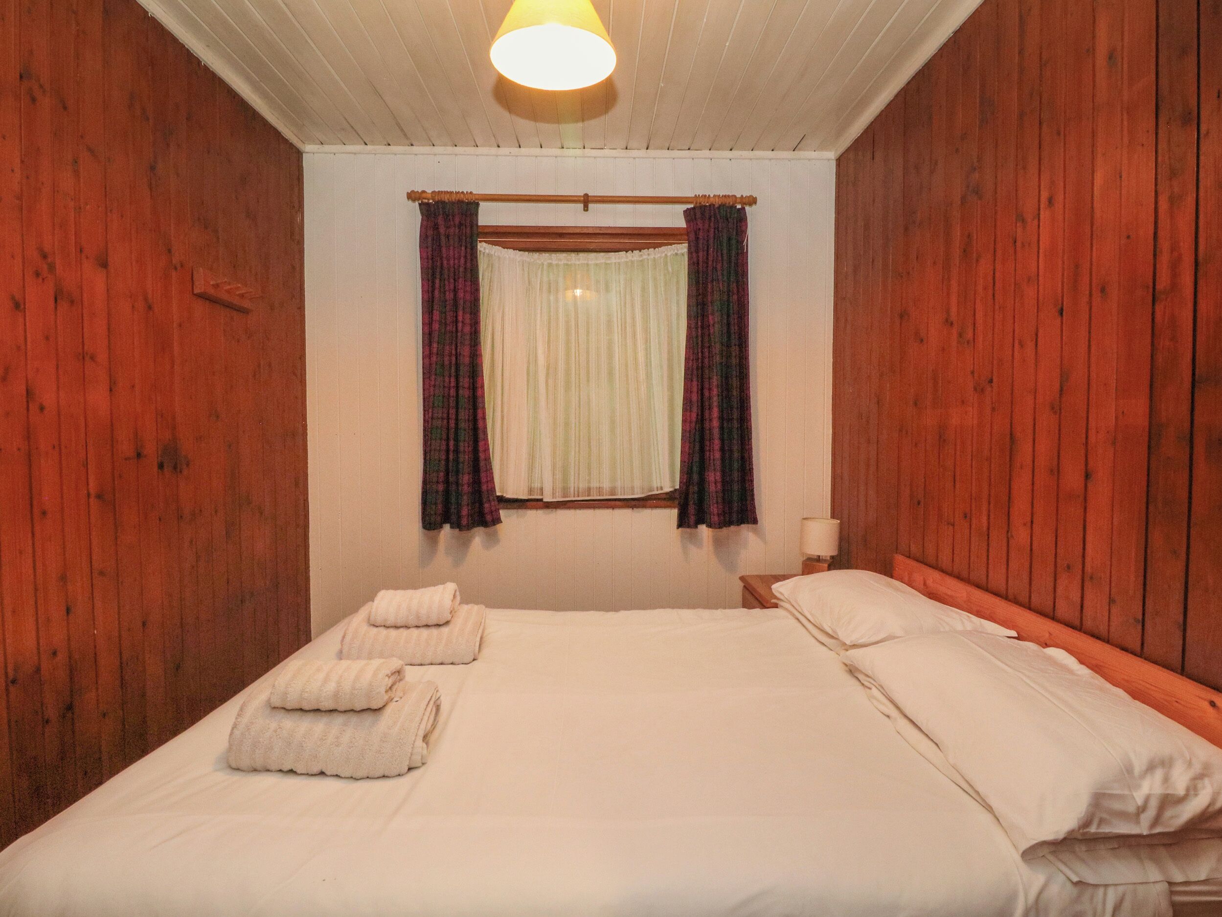 A room with wooden walls, a bed with white linens, a window with curtains, and a ceiling light.
