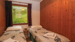 A room with two beds, each with a blanket featuring animal prints. There is a window with curtains, a lamp on a bedside table, and a view of greenery outside. - Lodge 5 - "hawthorn" (Isle of Arran)