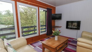 A living room with a sofa, a coffee table with a vase, a TV on the wall, and large windows with a view of trees. - Lodge 5 - "hawthorn" (Isle of Arran)