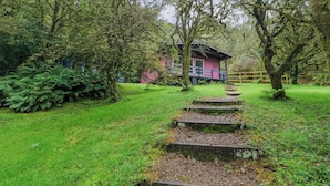 A pink building with a wooden deck, surrounded by lush greenery and a stone staircase leading up to it. - Lodge 5 - "hawthorn" (Isle of Arran)