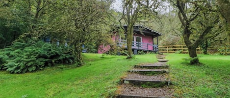 Un edificio rosa con una terraza de madera, rodeado de abundante vegetación y una escalera de piedra que conduce a él.