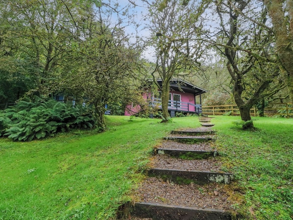 A pink building with a wooden deck, surrounded by lush greenery and a stone staircase leading up to it. - Lodge 5 - "hawthorn" (Isle of Arran)