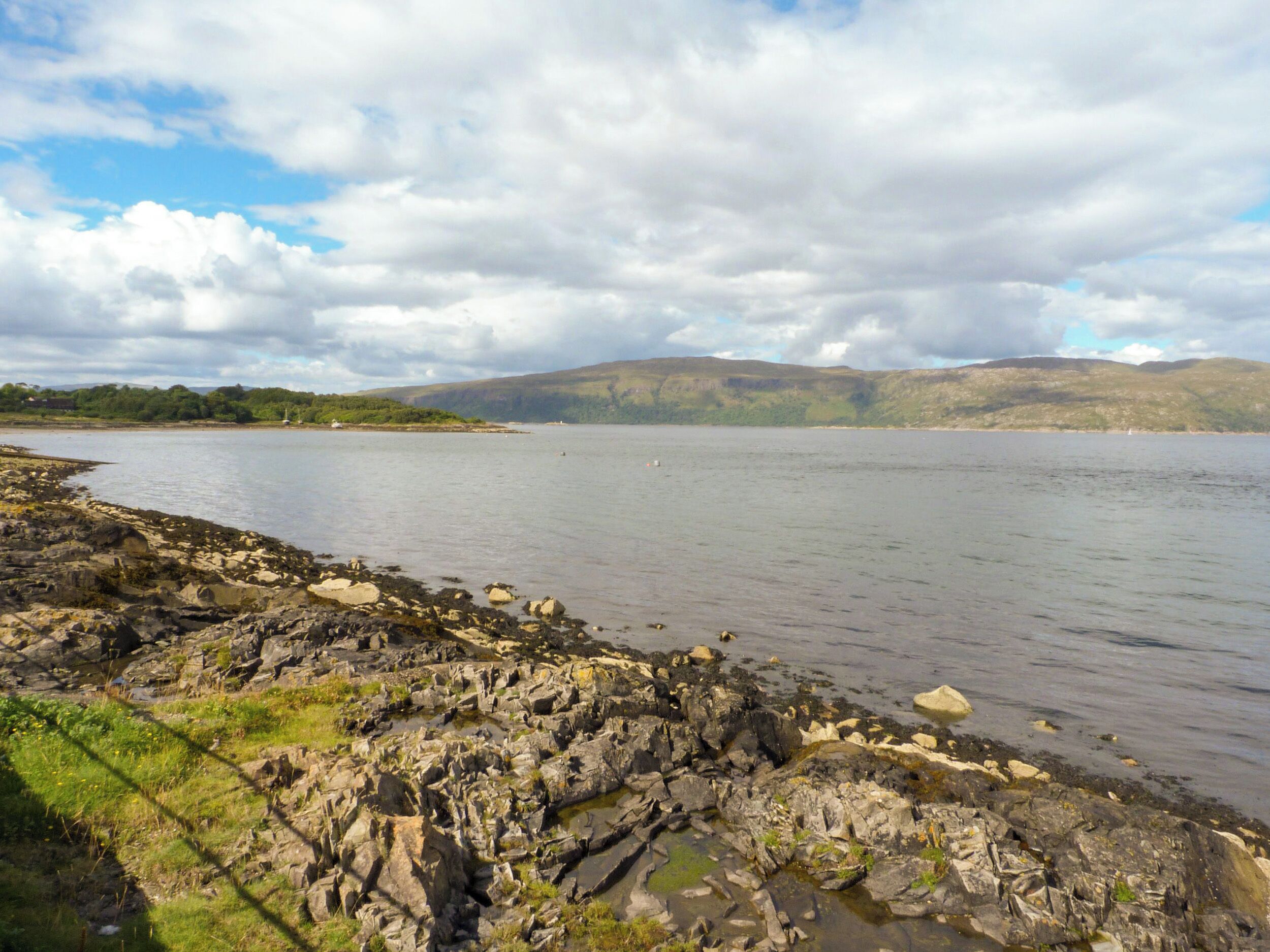A rocky shoreline with calm water, distant hills, and a partly cloudy sky.