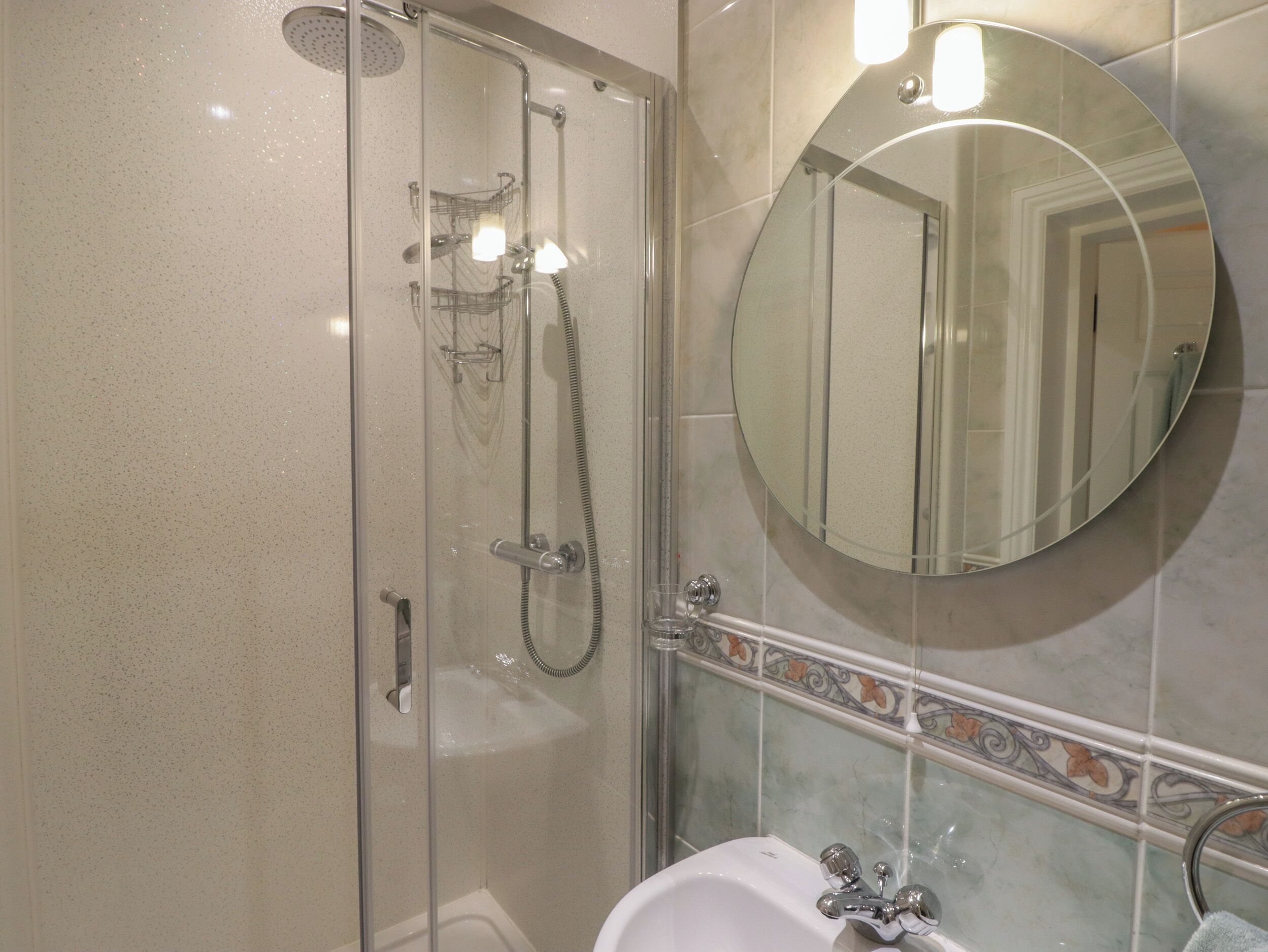 A bathroom with a glass-enclosed shower, a round mirror, and a sink with a decorative tile border.