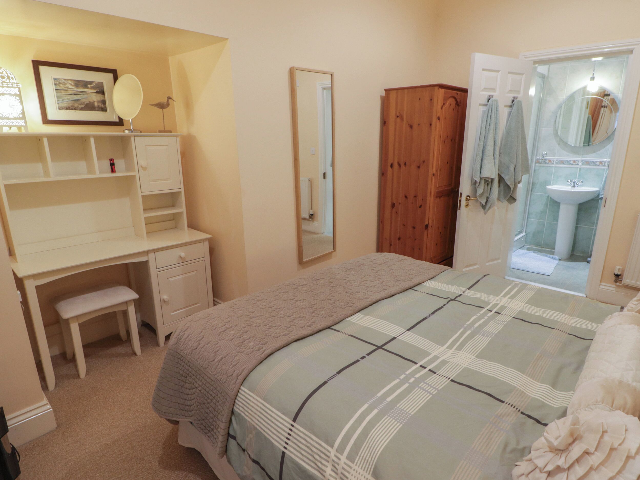 A bedroom with a bed, a desk, a wardrobe, a mirror, and a bathroom with a sink and towel rack.