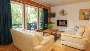A cozy living room with a beige sofa, a coffee table, and a TV. Large windows offer a view of the outdoors. - Lodge 4 - "birch" (Isle of Arran)