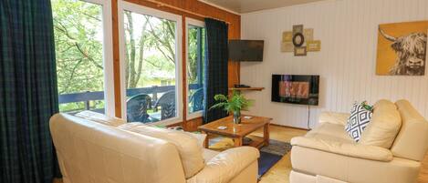 A cozy living room with a beige sofa, a coffee table, and a TV. Large windows offer a view of the outdoors.