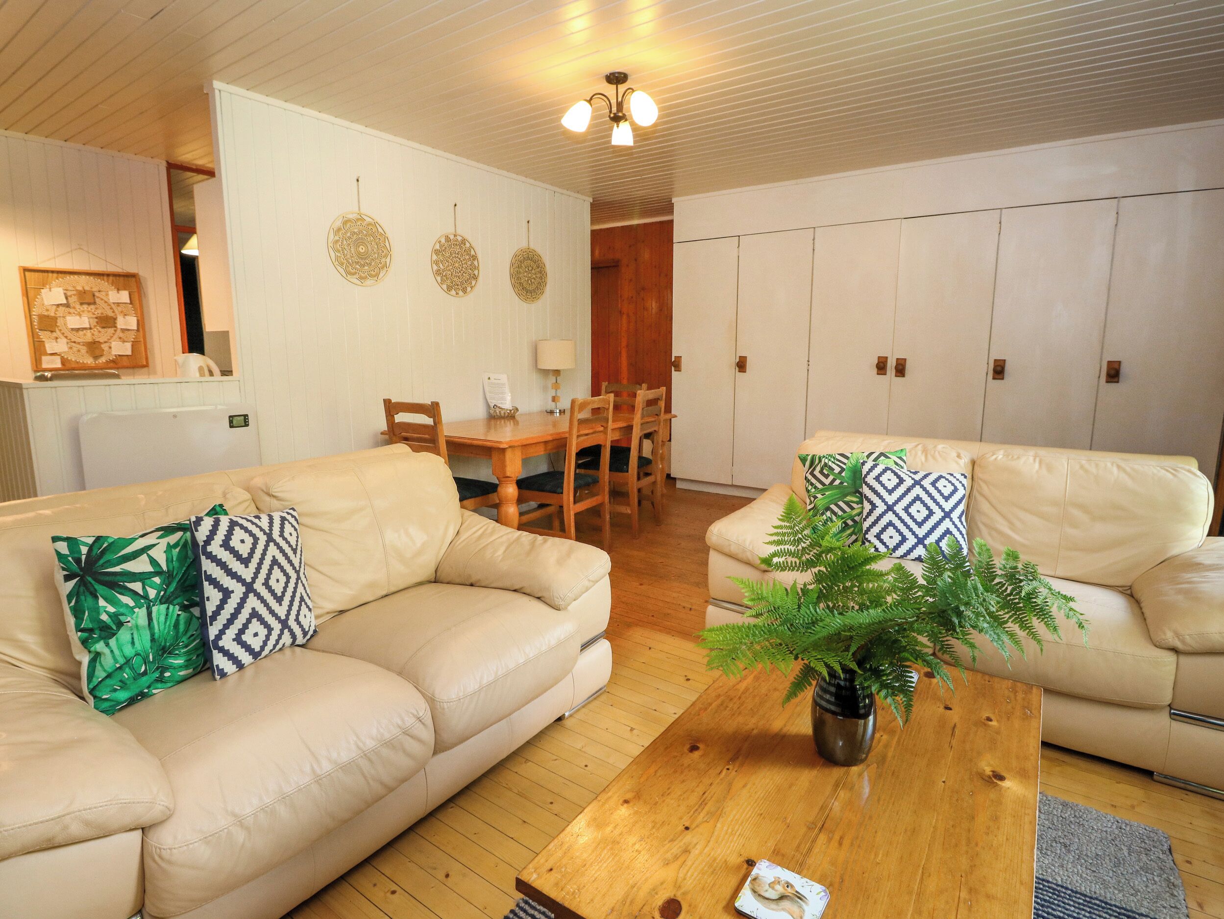 Una sala de estar con dos sofás color beige, una mesa de centro de madera con una planta, una mesa de comedor con sillas y un área de cocina al fondo.