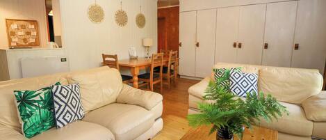 Una sala de estar con dos sofás color beige, una mesa de centro de madera con una planta, una mesa de comedor con sillas y un área de cocina al fondo.