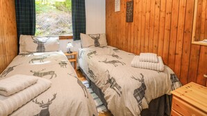 A room with two beds, each with a deer-themed duvet, a wooden nightstand, a window with plaid curtains, and a wooden wall. - Lodge 4 - "birch" (Isle of Arran)
