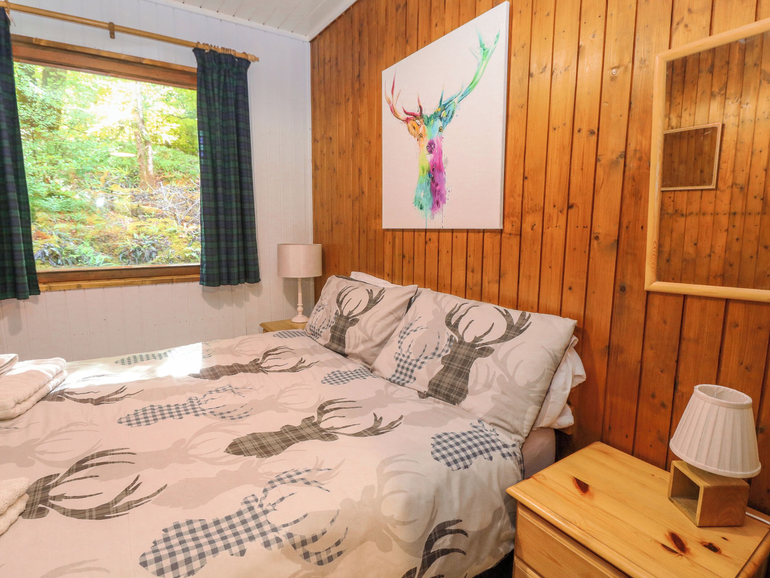 A bedroom with a bed featuring a deer pattern, a wooden headboard, a window with curtains, a bedside lamp, and a painting of a deer on the wall.