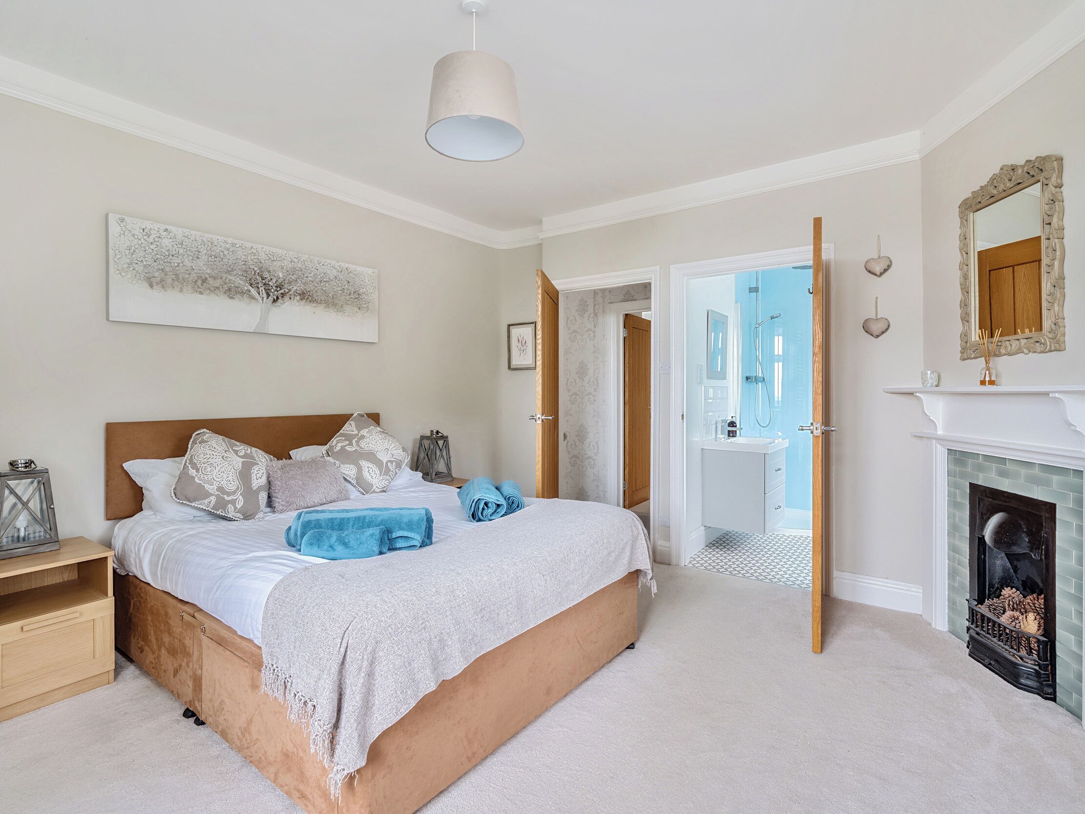 A bedroom with a large bed, a bedside table, a mirror, a fireplace, and an open door leading to a bathroom.