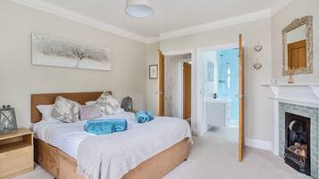 A bedroom with a large bed, a bedside table, a mirror, a fireplace, and an open door leading to a bathroom.