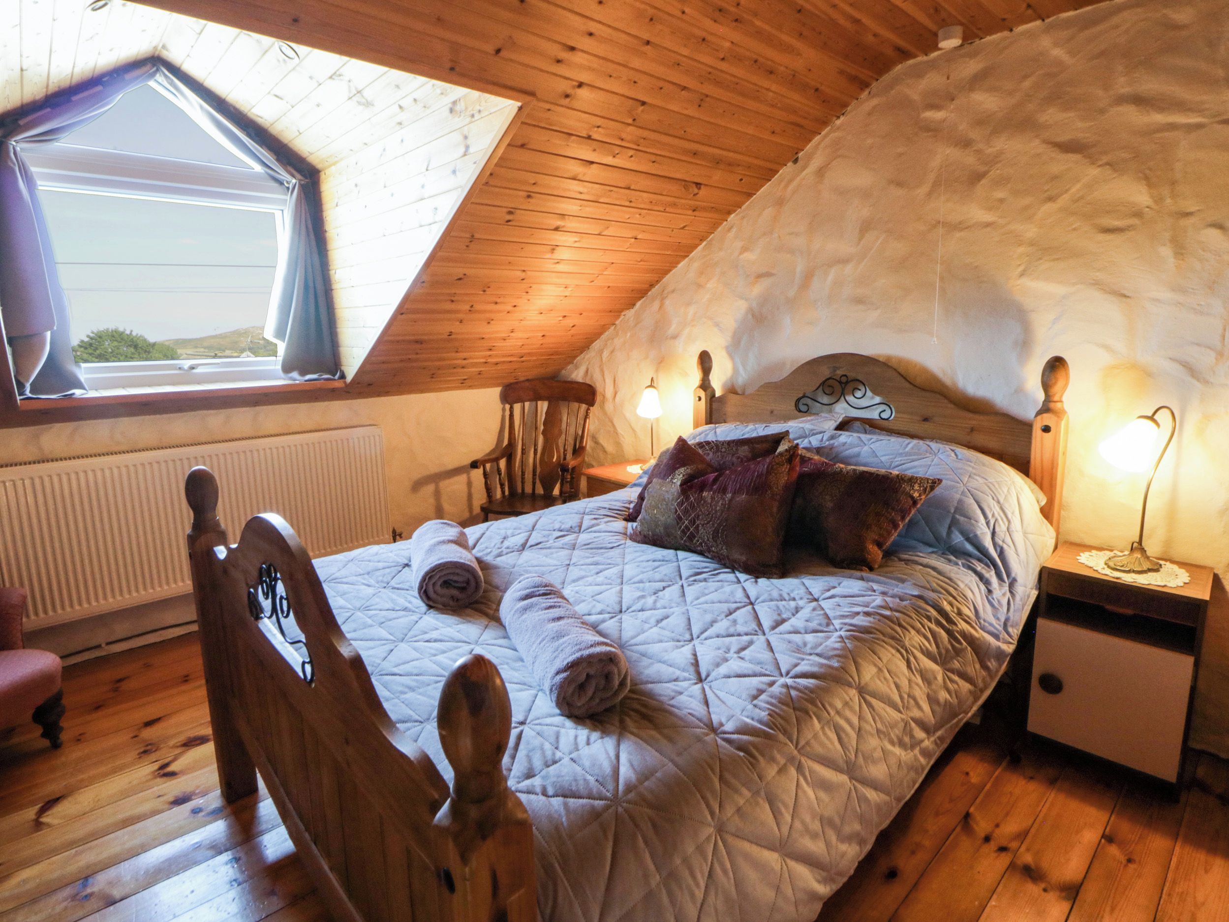 A bedroom with a sloped ceiling, a wooden bed with a headboard, a nightstand with a lamp, and a window with a view.