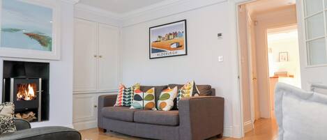 A living room with a fireplace, a grey sofa with colorful cushions, a framed picture of Aldeburgh, and a patterned rug.