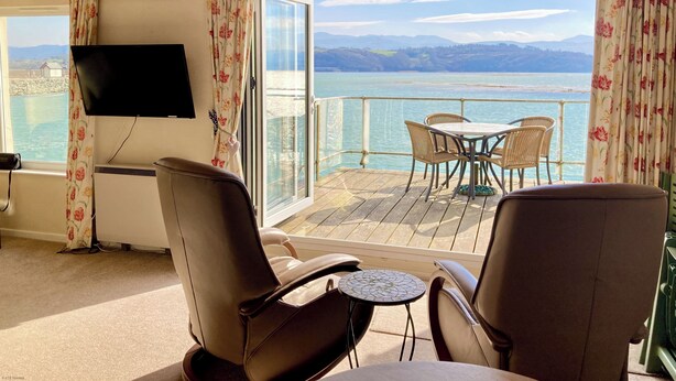 A room with a wooden ceiling, a television mounted on the wall, a window with floral curtains, a view of the sea, and a table with chairs. - 11 South Snowdon Wharf (Porthmadog)