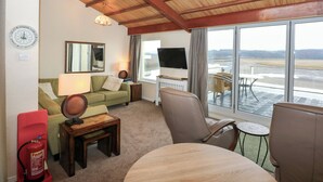 A living room with a wooden ceiling, a sofa, a coffee table, a TV, and a view of a landscape through large windows. - 11 South Snowdon Wharf (Porthmadog)