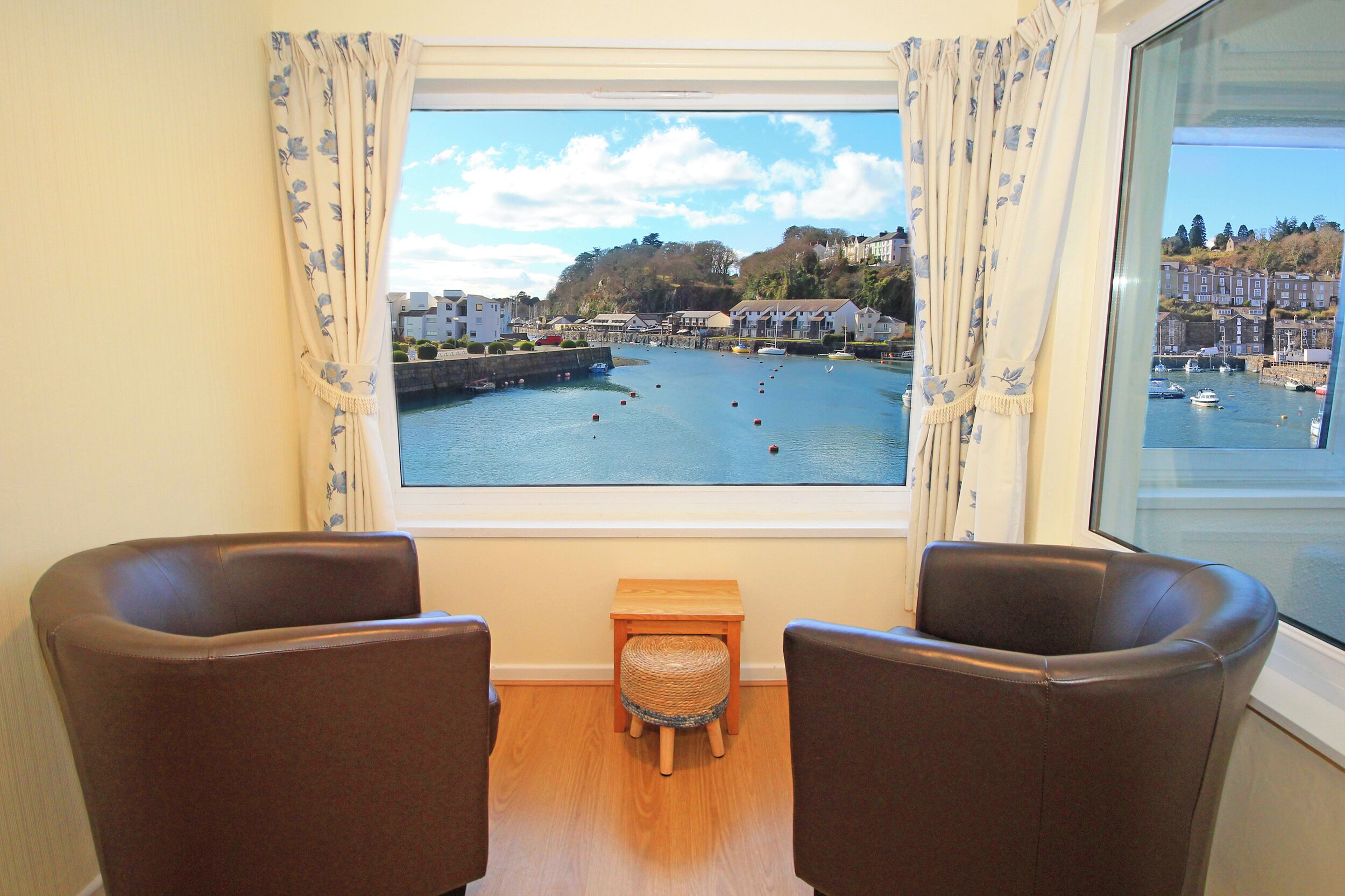Ein Zimmer mit zwei braunen Ledersesseln, einem kleinen Holzschemel und einem Fenster mit Blick auf einen Hafen und Gebäude.