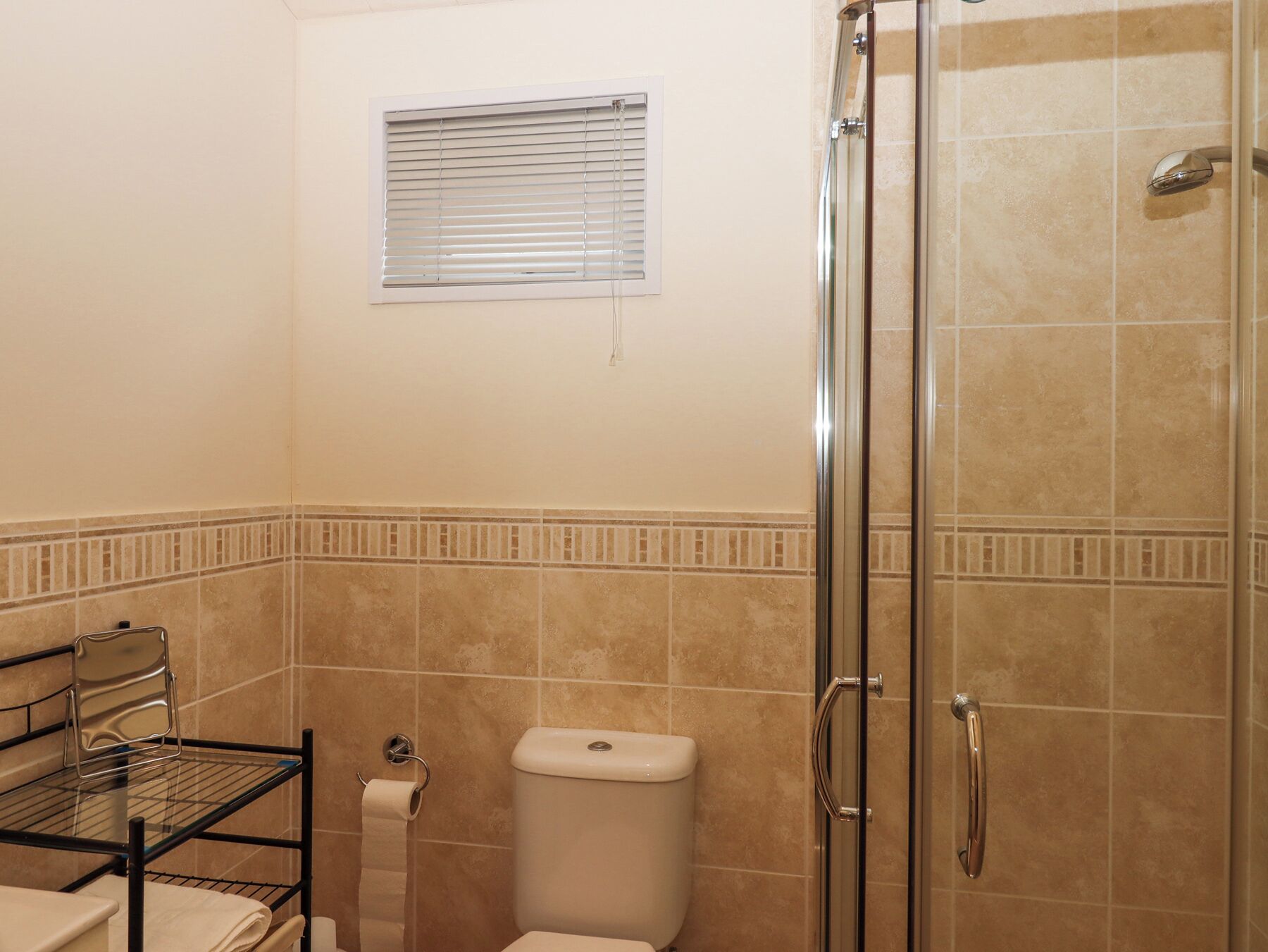 A bathroom with a glass shower enclosure, a toilet, a towel rack, and a window with blinds.