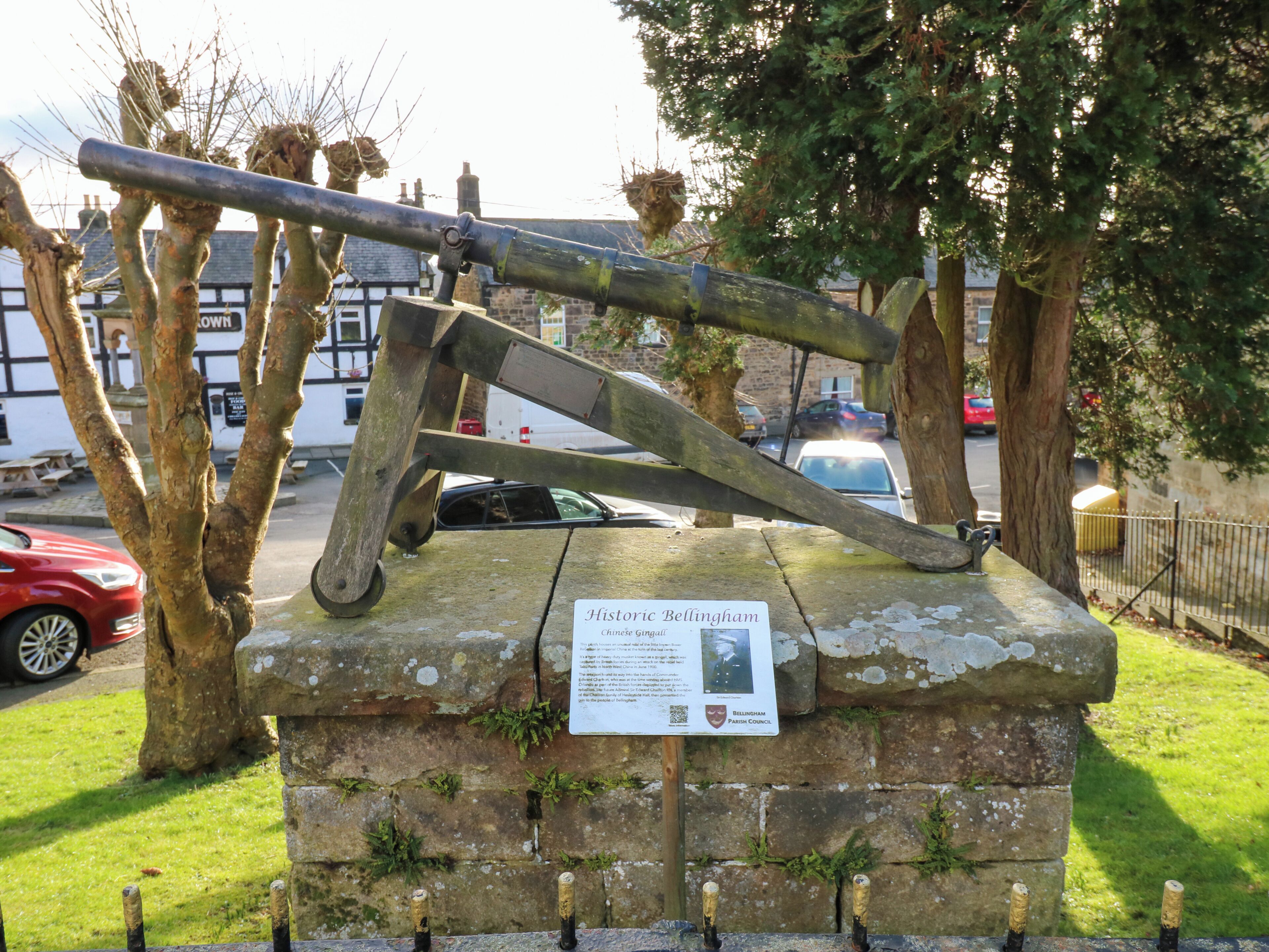 A historical stone monument with a plaque, surrounded by trees and a street view.