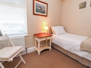 Een kleine, verzorgde slaapkamer met een enkel bed, een nachtkastje met lamp, een stoel en een lijst met een afbeelding aan de muur.