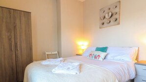 A neatly made bed with white linens and towels, a wooden wardrobe, a bedside table with a lamp, and a wall-mounted artwork. - Lytham House (Lytham St. Anne's)