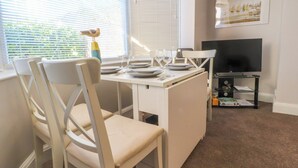 A dining area with a table set for four, white chairs, a TV on a stand, and a painting on the wall. - Lytham House (Lytham St. Anne's)