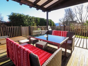 Een terras buiten met een tafel met glazen blad en rieten meubilair, omringd door een houten terras en een houten hek.
