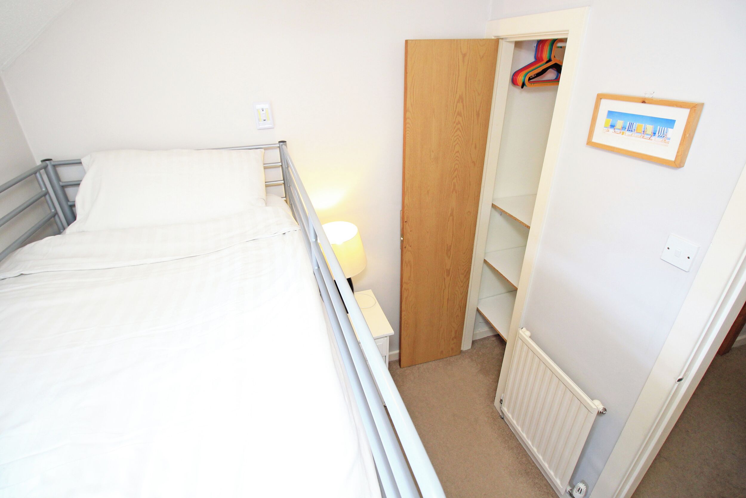 A single bed with a metal frame, a bedside table with a lamp, a wooden door leading to a closet, and a framed picture on the wall.