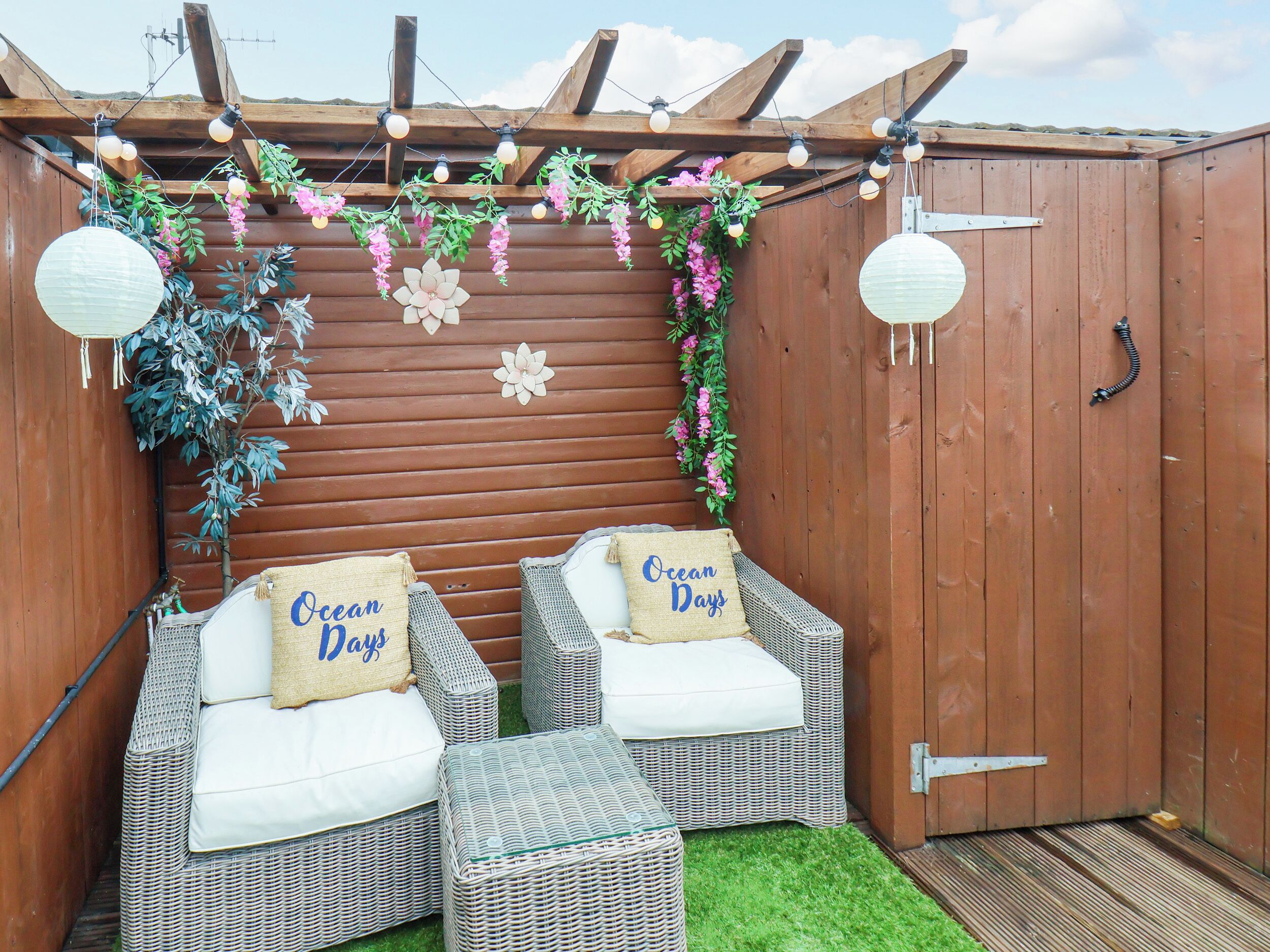 A cozy outdoor seating area with wicker furniture and a pergola overhead, decorated with lights and flowers.