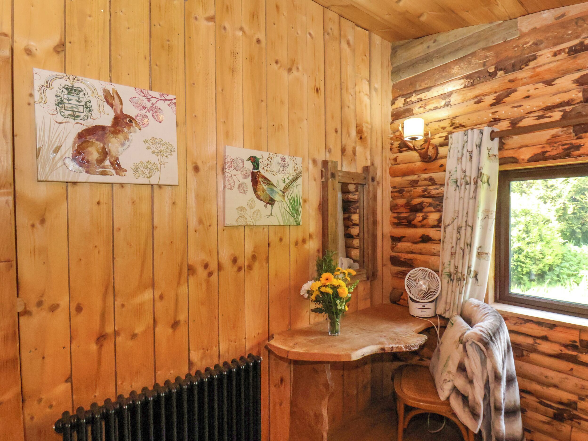 A wooden cabin interior with two framed paintings of birds, a small table with a vase of flowers, a chair, and a window with curtains.