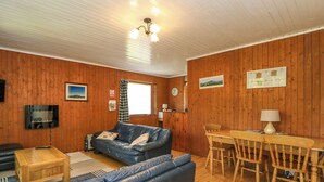 A cozy room with a wooden floor, a blue leather sofa, a wooden dining table with chairs, a television mounted on the wall, and a ceiling fan. - Lodge 6 - "alder" (Isle of Arran)