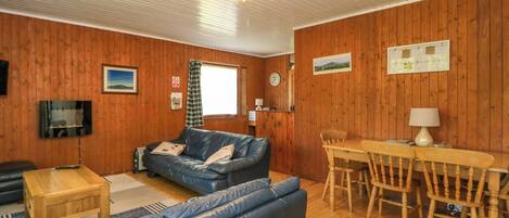 Een knus interieur met een houten vloer, een blauwe leren bank, een houten eettafel met stoelen, een aan de muur bevestigde televisie en een plafondventilator.