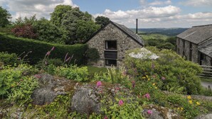 Un edificio de piedra con una ventana pequeña y una puerta, rodeado de abundante follaje y plantas en flor.
