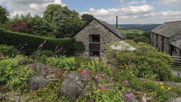 Un edificio de piedra con una ventana pequeña y una puerta, rodeado de abundante follaje y plantas en flor.