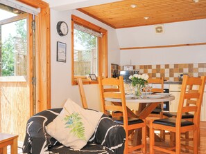 A cozy dining area with wooden chairs and a table set for two, a clock on the wall, and a window with blinds. - Lodge 47 - Primrose Lodge (Newton Abbot)