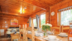 A wooden cabin interior with a dining table set for four, wooden chairs, a vase of white flowers, and a clock on the wall. - Lodge 32 - Cowslip (Newton Abbot)