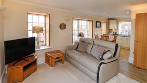 A living room with a sofa, TV, and wooden side tables. There are windows and a view of the outside.