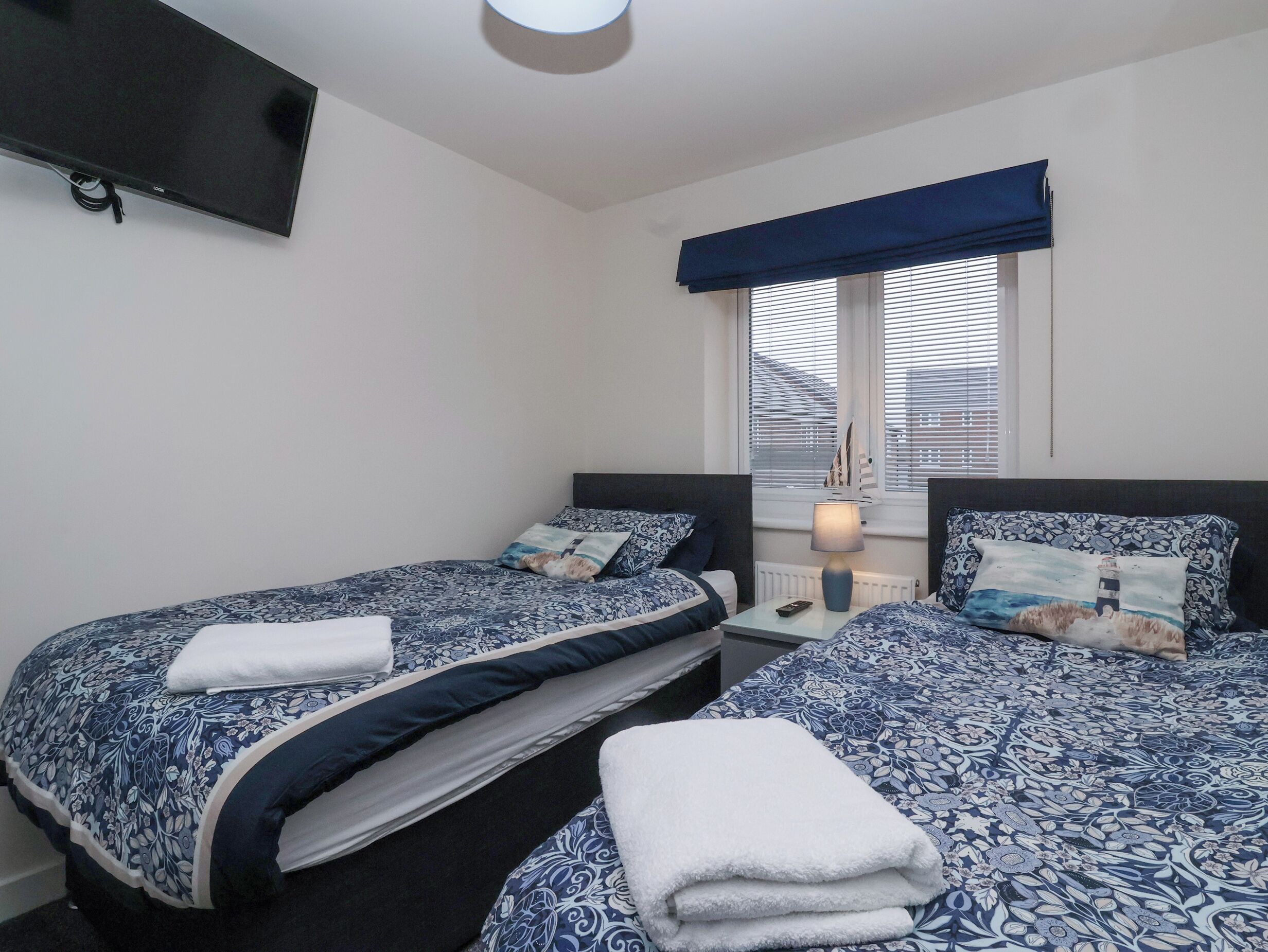 Un dormitorio con dos camas, un televisor montado en la pared, una ventana con persianas y una lámpara de noche.