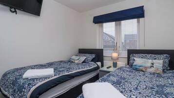 Un dormitorio con dos camas, un televisor montado en la pared, una ventana con persianas y una lámpara de noche.