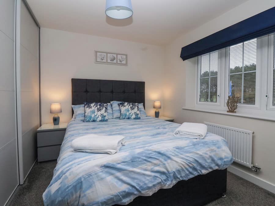 A neatly made bed with a blue and white patterned bedspread, two bedside lamps, a headboard, and a window with blinds.