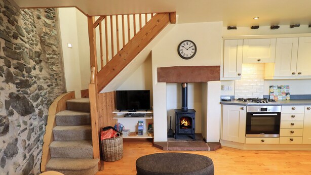 A cozy living room with a stone wall, a wooden staircase, a fireplace, a television, and a kitchen area with cabinets and a stove. - Squirrel Cottage (Keswick)