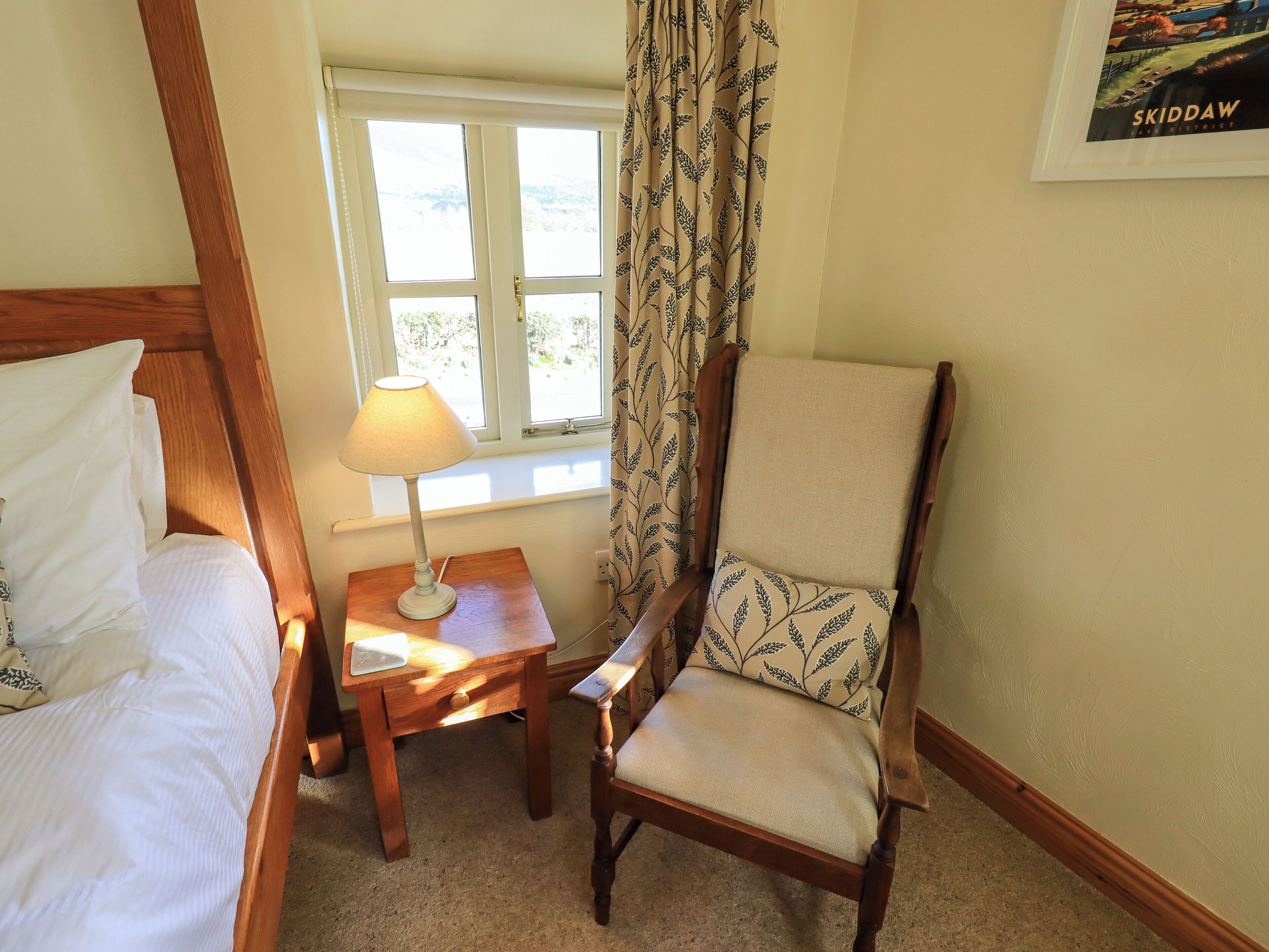 A bedroom with a bed, a wooden nightstand with a lamp, a chair with a patterned cushion, and a window with curtains.