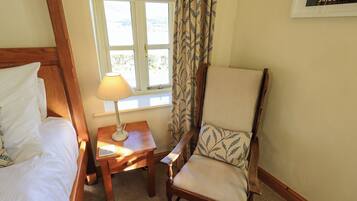 A bedroom with a bed, a wooden nightstand with a lamp, a chair with a patterned cushion, and a window with curtains.